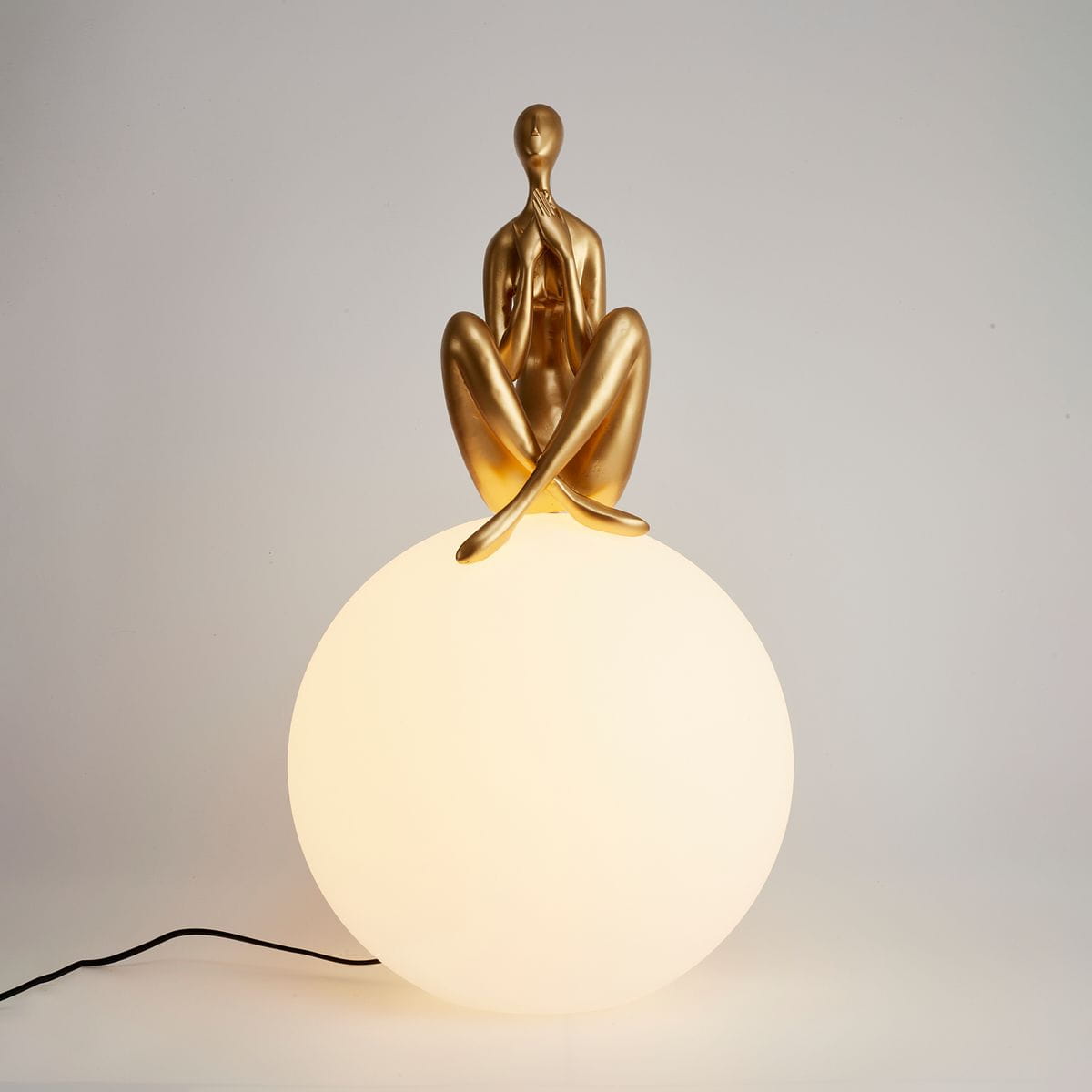 Lampa stołowa Woman-3 z figurką w złotym kolorze siedzącą na białej, okrągłej podstawie. Produkt prezentowany frontalnie, z widocznym kablem zasilającym. Idealna do nowoczesnych wnętrz jako dekoracyjny element oświetlenia.