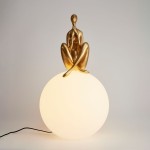 Lampa stołowa Woman-3 z figurką w złotym kolorze siedzącą na białej, okrągłej podstawie. Produkt prezentowany frontalnie, z widocznym kablem zasilającym. Idealna do nowoczesnych wnętrz jako dekoracyjny element oświetlenia.