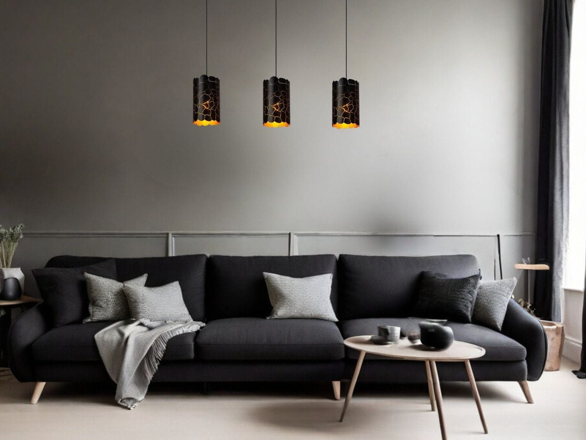 Czarna lampa wisząca Almelo z trzema oprawami E27, o cylindrycznym kształcie i dekoracyjnym wzorze, zawieszona nad czarną sofą. Tło w stonowanej szarości, minimalistyczne otoczenie z drewnianym stolikiem i poduszkami.