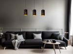 Czarna lampa wisząca Almelo z trzema oprawami E27, o cylindrycznym kształcie i dekoracyjnym wzorze, zawieszona nad czarną sofą. Tło w stonowanej szarości, minimalistyczne otoczenie z drewnianym stolikiem i poduszkami.