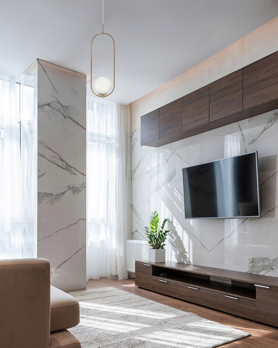 Lampa wisząca Ritus 1xE14 w złotej oprawie z białym kloszem, zawieszona w nowoczesnym wnętrzu. Widoczna w tle ściana z marmurowym wzorem, telewizor na drewnianej szafce, roślinność w donicy.