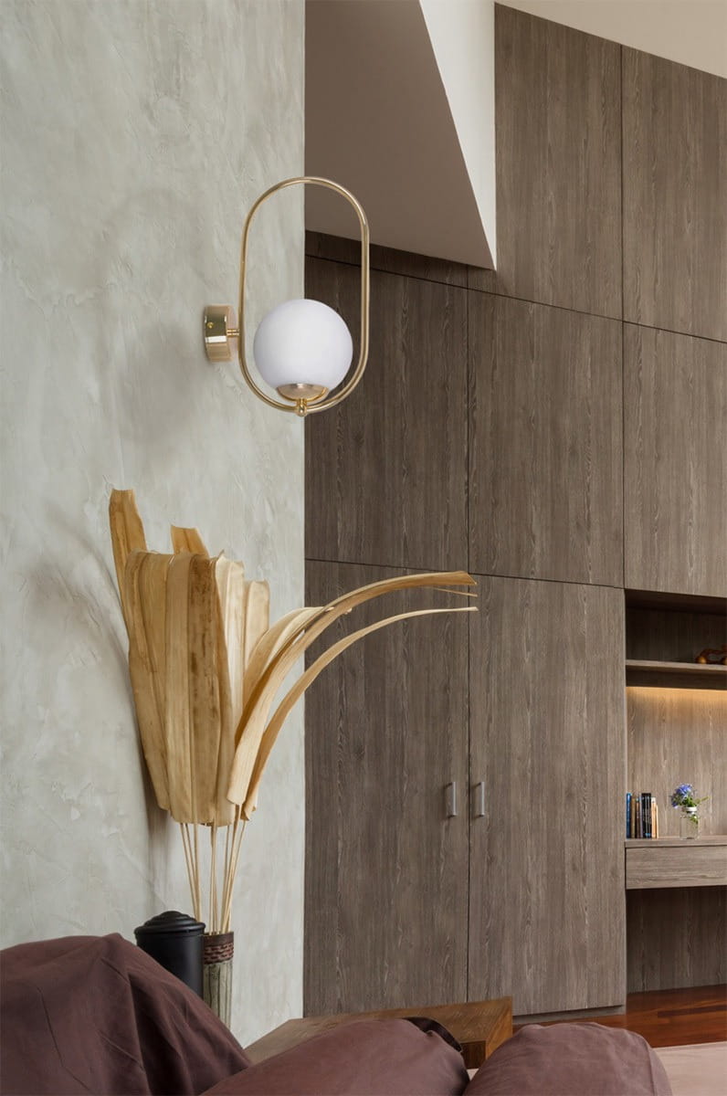 Kinkiet Ritus 1xE14 w kolorze złotym i białym, zamontowany na ścianie. Owalna, biała kula ze szkła otoczona złotą ramą. W tle widoczne elementy drewniane oraz rośliny dekoracyjne, co tworzy nowoczesny wystrój wnętrza.