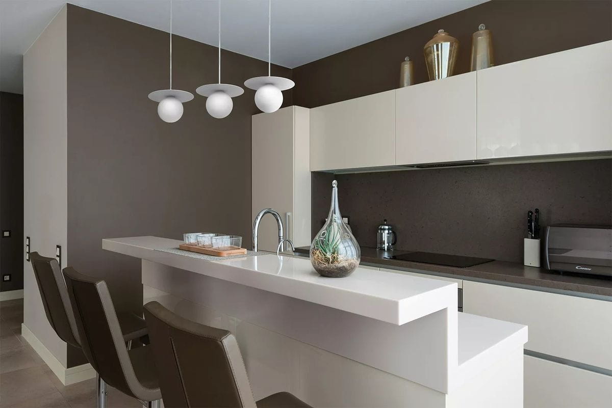 Lampa wisząca Robin D18 w białym kolorze z jednym gniazdem G9, zawieszona nad białym blatem kuchennym. Kształt okrągłych kloszy z gładką powierzchnią, w nowoczesnym wnętrzu z ciemnobrązowymi ścianami i minimalistycznymi dodatkami.