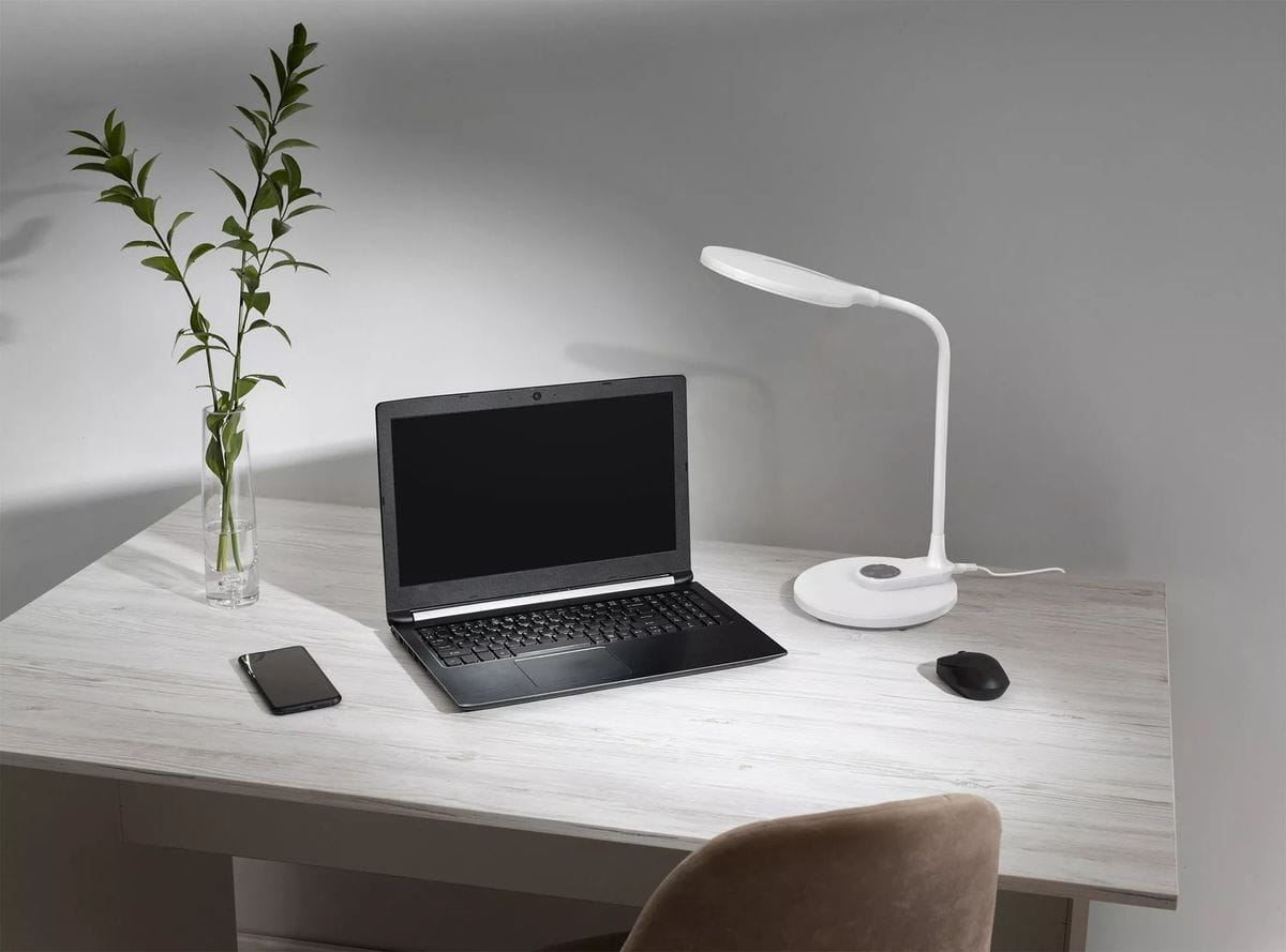 Lampka biurkowa Gaja w kolorze białym, z jedną diodą LED o mocy 9W i regulowanej temperaturze barwowej 3000-5700K. Ustawiona na biurku obok laptopa i telefonu, z wygodnym kształtem oraz podstawą okrągłą. W tle widoczna roślina w szklanym wazonie.