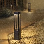 Czarna lampa ogrodowa Nutri H60 z jedną diodą LED 10W, ustawiona na podłożu z kamieni. Lampa ma prostokątny kształt, z przezroczystym panelem u góry, emitującym światło. Tło przedstawia deszczową scenerię w ogrodzie.