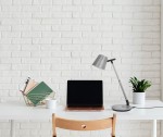 Lampka biurkowa Bolek w kolorze grafitowym z regulowanym ramieniem, z jedną żarówką LED 7W o temperaturze barwowej 3000-6000K, umieszczona na białym biurku obok laptopa, filiżanki i rośliny doniczkowej, na tle białej ceglanej ściany.