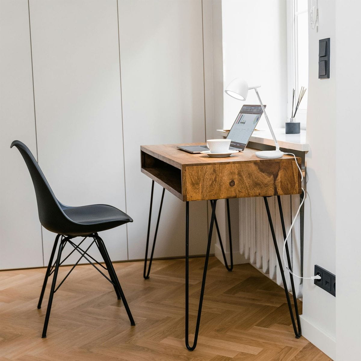 Lampka biurkowa Lolek w białym kolorze, z regulowanym ramieniem, umieszczona obok laptopa na drewnianym biurku. W tle czarna, ergonomiczna krzesło i okno. Oświetlenie LED o zmiennej temperaturze barwowej 3000-6500K.