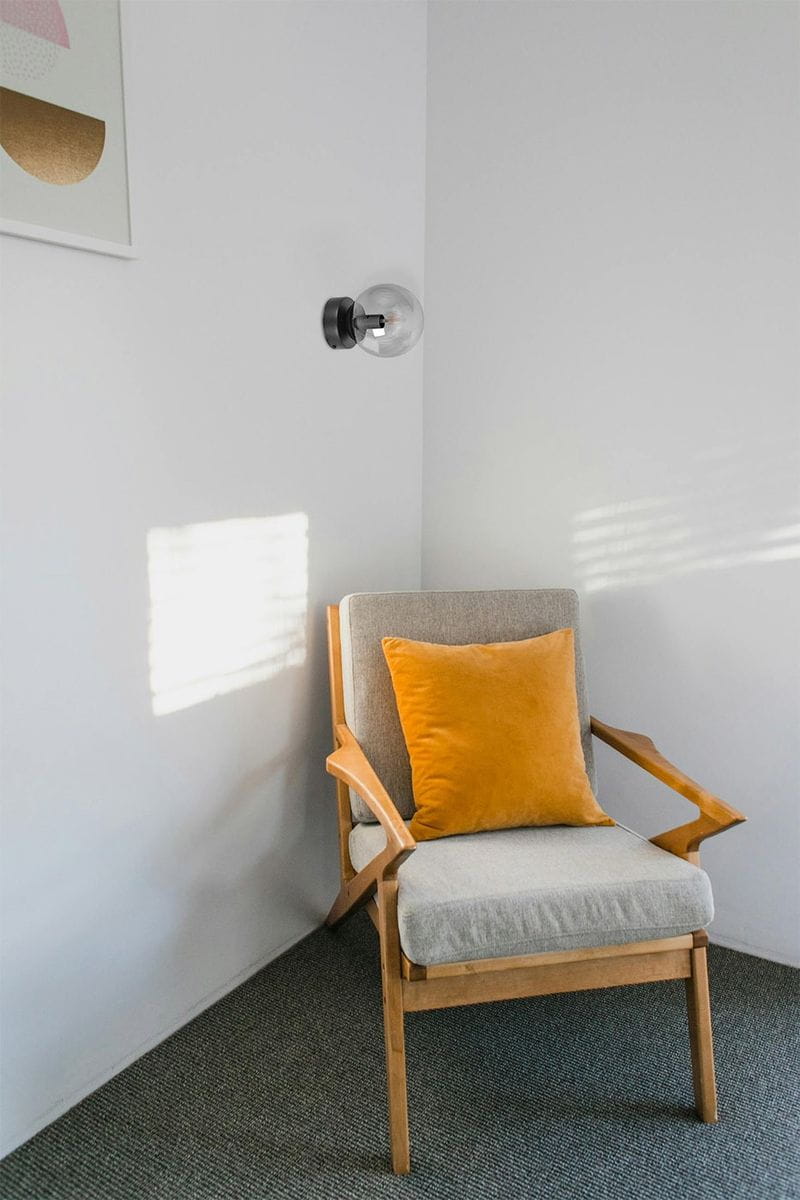 Czarna i grafitowa lampa sufitowa Megan z miejscem na żarówkę E14, zamontowana na ścianie w rogu pokoju. W tle widoczny szary fotel z pomarańczową poduszką, a całość utrzymana w jasnej, minimalistycznej aranżacji.