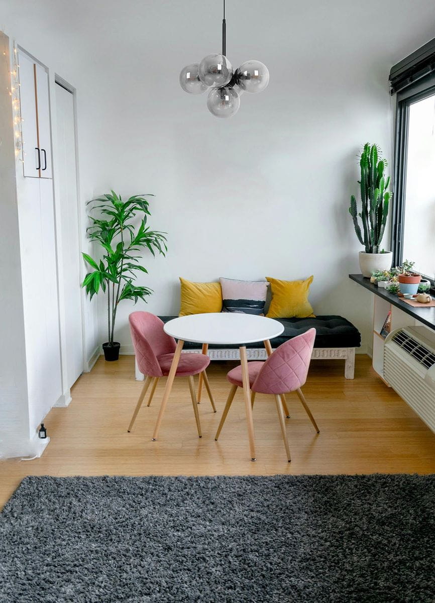 Czarna lampa wisząca Megan z pięcioma źródłami światła E14, zawieszona na cienkim kablu. Szklane klosze w odcieniu grafitowym, tworzą nowoczesny wygląd. Lampa umieszczona w jasnym wnętrzu z roślinami i meblami w odcieniach różu i żółci.