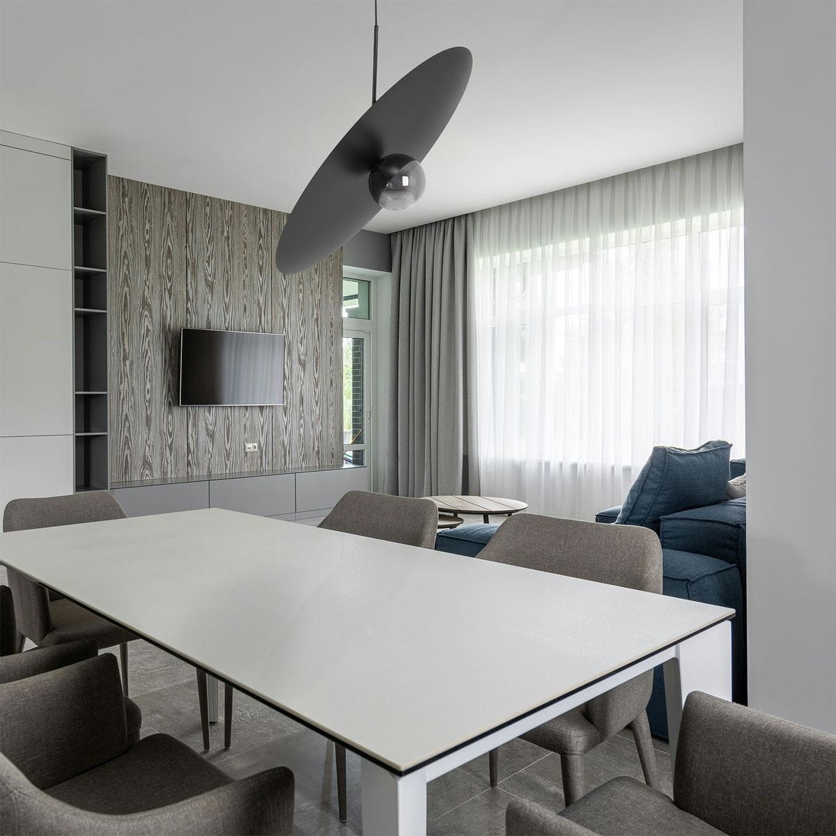 Lampa wisząca Fermi D60 w kolorze czarnym i grafitowym, z jednym źródłem światła G9. Zawieszona nad białym stołem w nowoczesnym wnętrzu, w otoczeniu szarych krzeseł, z widokiem na zasłony i ścianę z telewizorem.