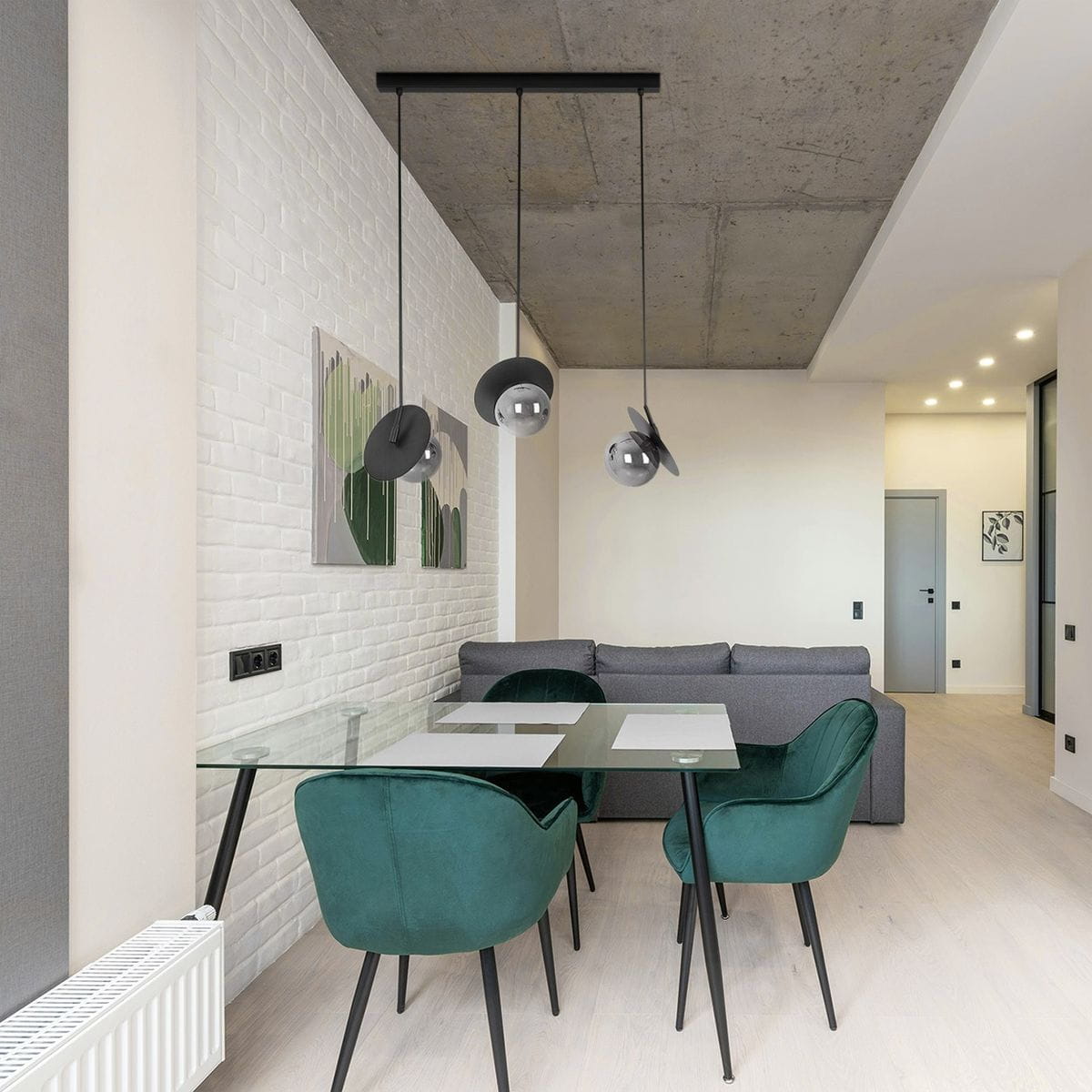 Lampa wisząca Fermi w kolorze czarnym i grafitowym z trzema kloszami z metalowymi elementami. Zawieszona na długich linkach nad szklanym stołem w nowoczesnej aranżacji jadalni, z zielonymi krzesłami i jasnym tłem.