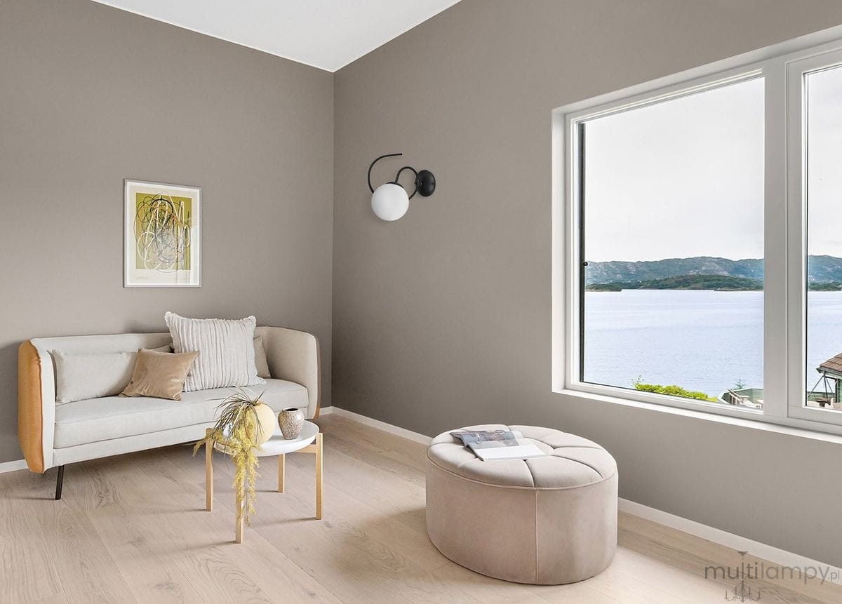 Kinkiet Nela w kolorze czarnym i białym, zamontowany na szarej ścianie. Oświetlenie ma okrągłą, białą oprawę i minimalistyczny, nowoczesny design. W tle widoczna jasna kanapa z poduszkami oraz okrągła pufa.