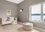 Kinkiet Nela w kolorze czarnym i białym, zamontowany na szarej ścianie. Oświetlenie ma okrągłą, białą oprawę i minimalistyczny, nowoczesny design. W tle widoczna jasna kanapa z poduszkami oraz okrągła pufa.