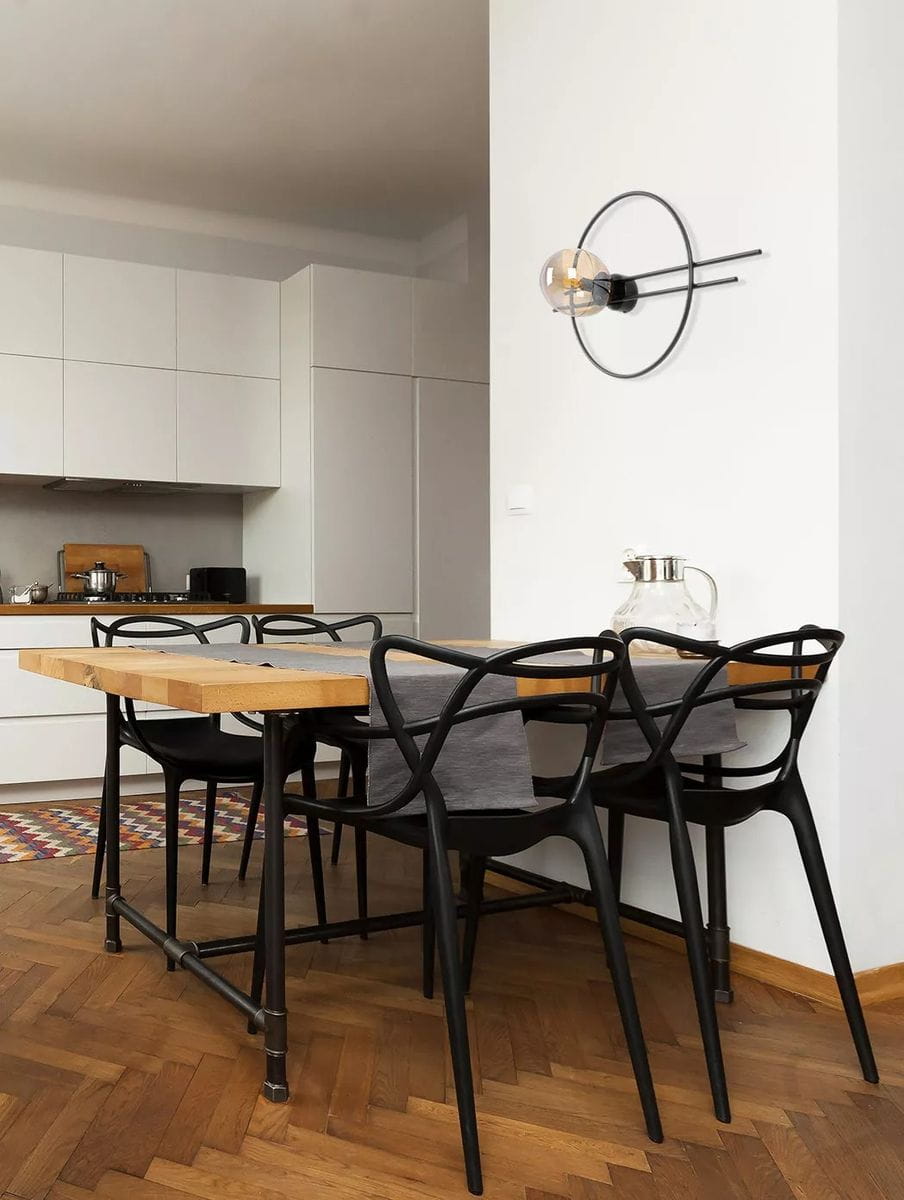 Kinkiet Corsa 1xE14 w kolorze czarnym z bursztynową żarówką, zamontowany na białej ścianie. Produkt ma minimalistyczny, okrągły kształt z metalowymi elementami. Widoczny w kontekście nowoczesnej kuchni z drewnianym stołem i czarnymi krzesłami.