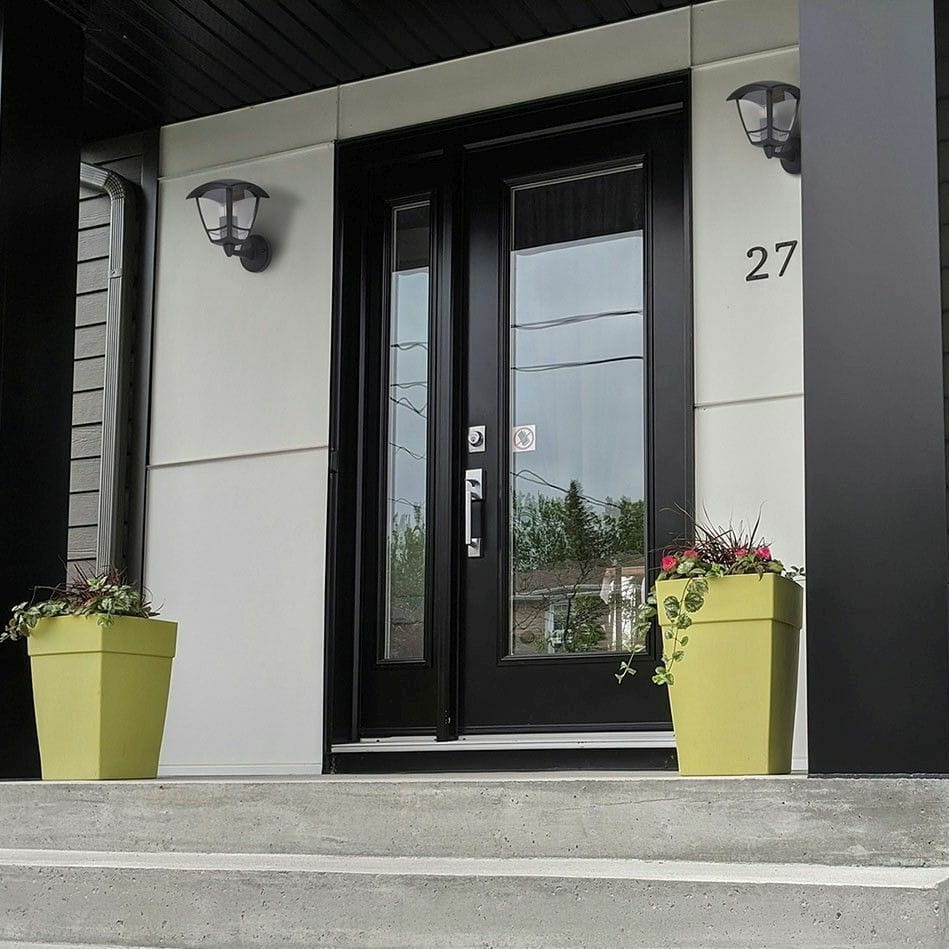 Kinkiet elewacyjny Napoli w kolorze czarnym, z jedną oprawą na żarówkę E27, zamontowany obok drzwi wejściowych. Stylowy, metalowy design z przezroczystym kloszem, umieszczony na ścianie w aranżacji z zielonymi donicami.