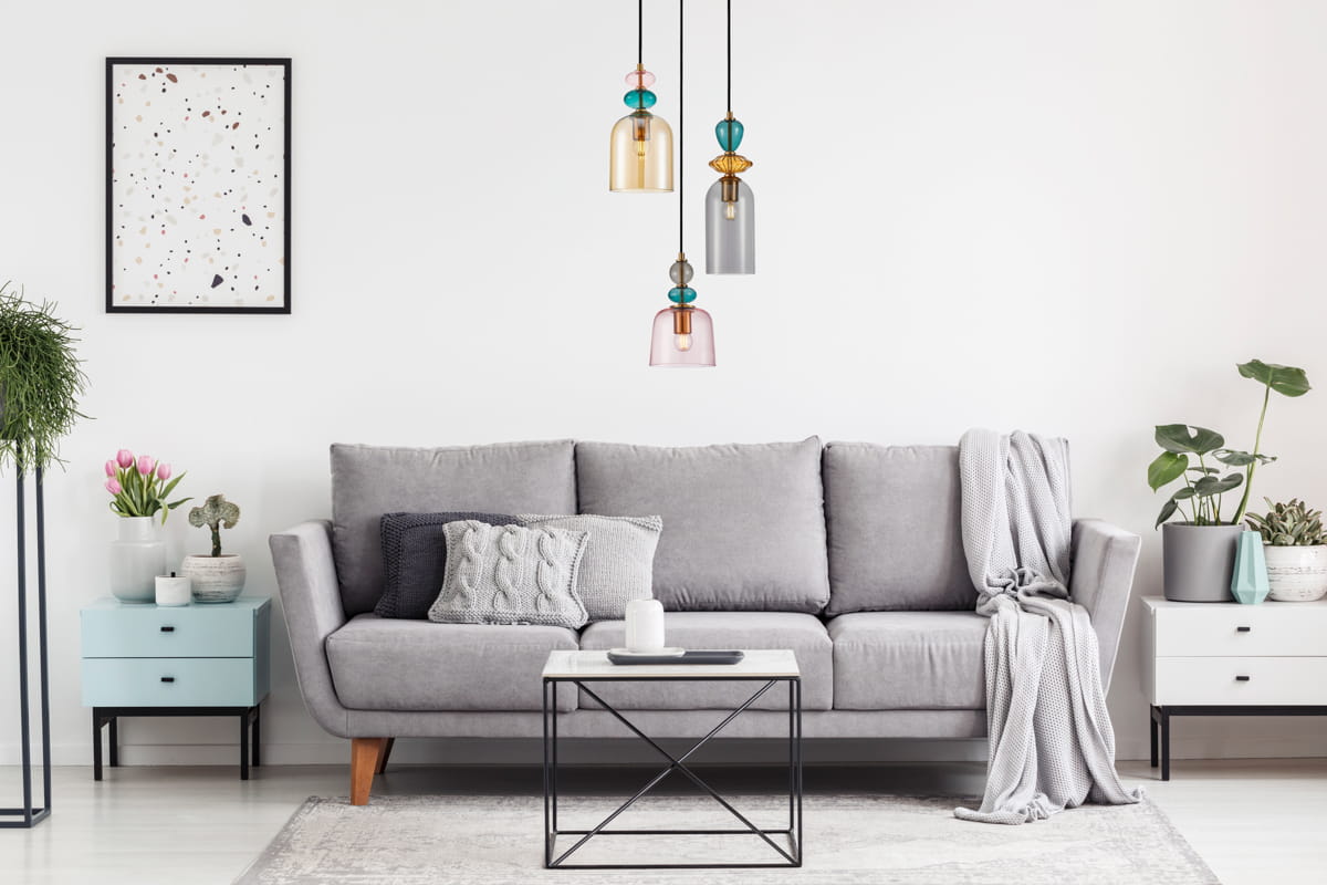 Lampa wisząca Namos 3xE27 w kolorze czarnym, z trzema kloszami w różnych kolorach (żółtym, szarym, różowym), zawieszona nad szarym sofą w nowoczesnym wnętrzu. Długie kable w czarnym kolorze. Tło jasne, minimalistyczne z roślinami i dekoracjami.