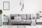 Lampa wisząca Namos 3xE27 w kolorze czarnym, z trzema kloszami w różnych kolorach (żółtym, szarym, różowym), zawieszona nad szarym sofą w nowoczesnym wnętrzu. Długie kable w czarnym kolorze. Tło jasne, minimalistyczne z roślinami i dekoracjami.