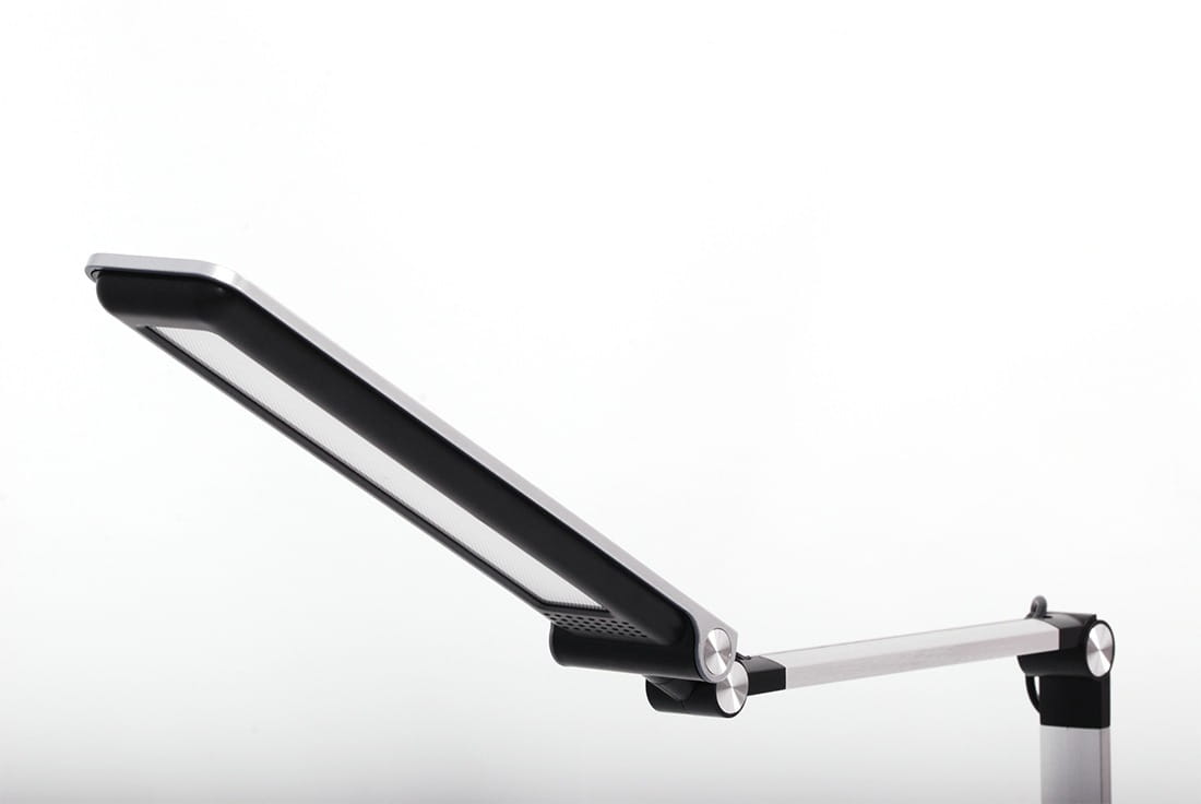 Srebrna lampka biurkowa Rico z jedną diodą LED (10W), z regulowanym ramieniem. Ujęcie z boku pokazuje panel świetlny z czarną ramką oraz metalowe elementy mocujące. Tło białe, co podkreśla nowoczesny design lampki.