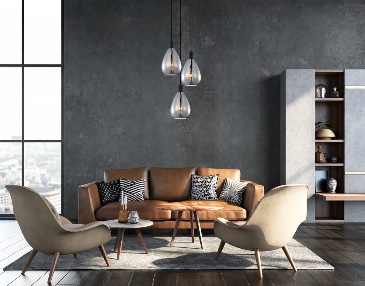 Lampa wisząca Zulo z trzema żarówkami E27, w kolorze czarnym z dymionymi kloszami, zawieszona w nowoczesnym wnętrzu. W tle skórzana sofa, okrągły stół oraz designerskie fotele na drewnianych nogach.