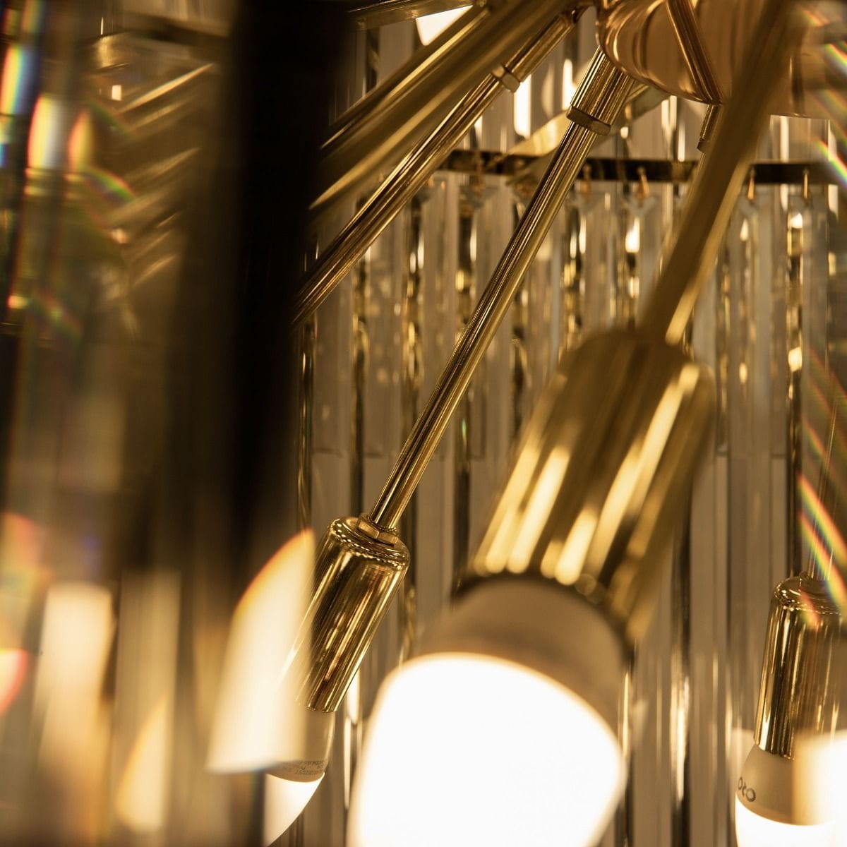 Luxe żyrandol w złotym kolorze, z sześcioma mocowaniami na żarówki E14. Ujęcie z bliska na metalowe elementy i przezroczyste kryształy, które odbijają światło. Widać detale wykończenia i stylowy design.