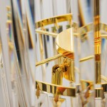 Żyrandol Luxe 40xE14 w odcieniu złotym. Widok zbliżony na detale, w tym złotą podstawę oraz przezroczyste elementy kryształowe, podkreślające elegancki design. Produkt zawiera mocowanie oraz dekoracyjne frędzle.