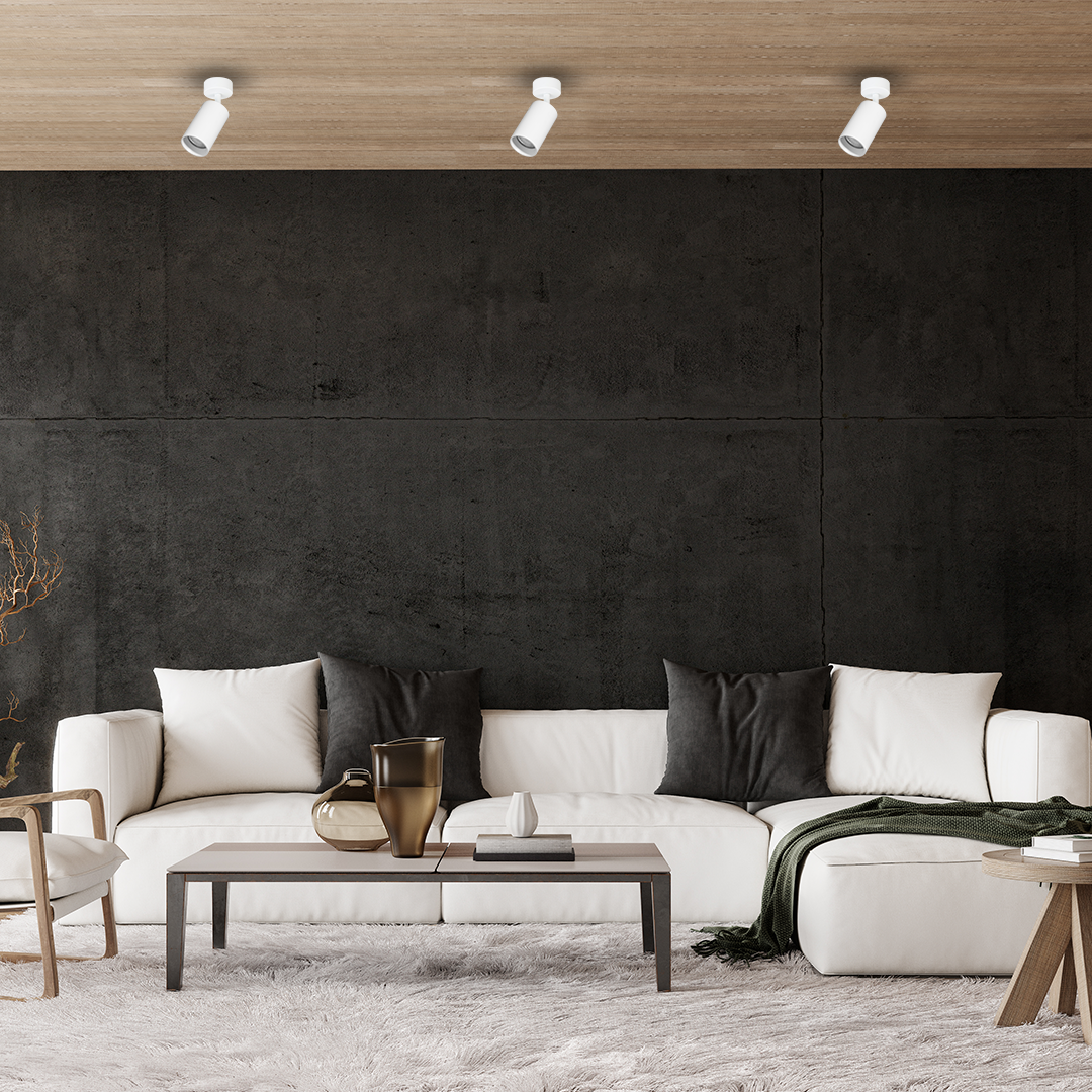 Lampa sufitowa typu spot w kolorze białym, z zamocowaniem na suficie, z jedną oprawą GU10. Ujęcie z góry, pokazujące lampę w nowoczesnym wnętrzu z ciemną ścianą i jasnymi meblami.