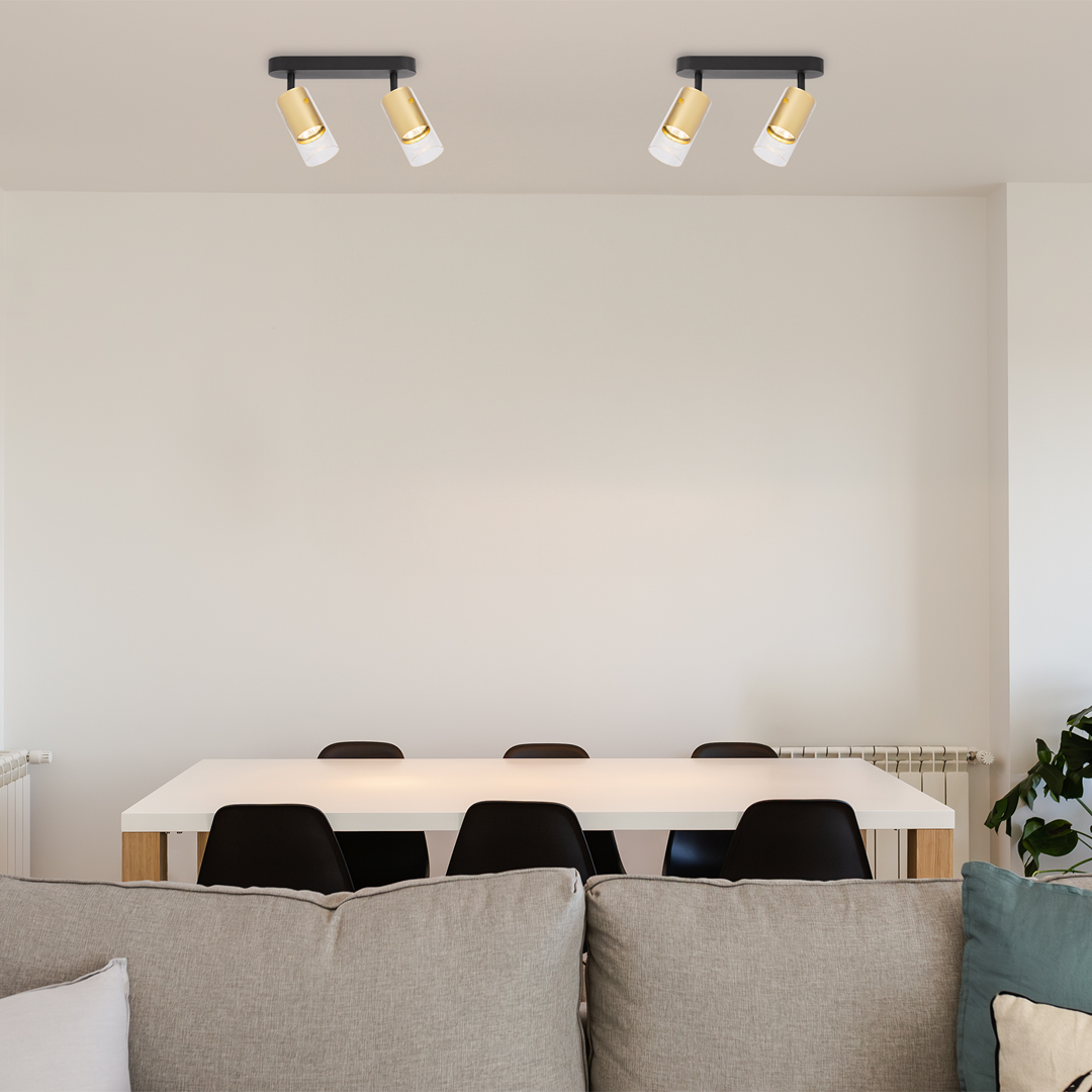 Czarna lampa sufitowa Ricky z dwoma spotami GU10, z metalowymi złotymi detalami, zamontowana na prostokątnej podstawie. Ujęcie z dołu, pokazujące lampę nad białym stołem i czarnymi krzesłami w nowoczesnym wnętrzu.
