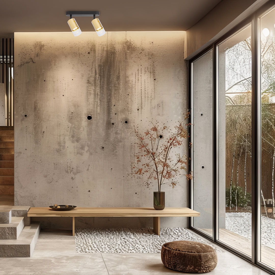 Czarna lampa sufitowa typu spot z dwoma żarówkami GU10, zamocowana na metalowej podstawie. Obudowy żarówek w kolorze złotym. Lampa umieszczona w nowoczesnym wnętrzu z betonową ścianą i drewnianym meblem.