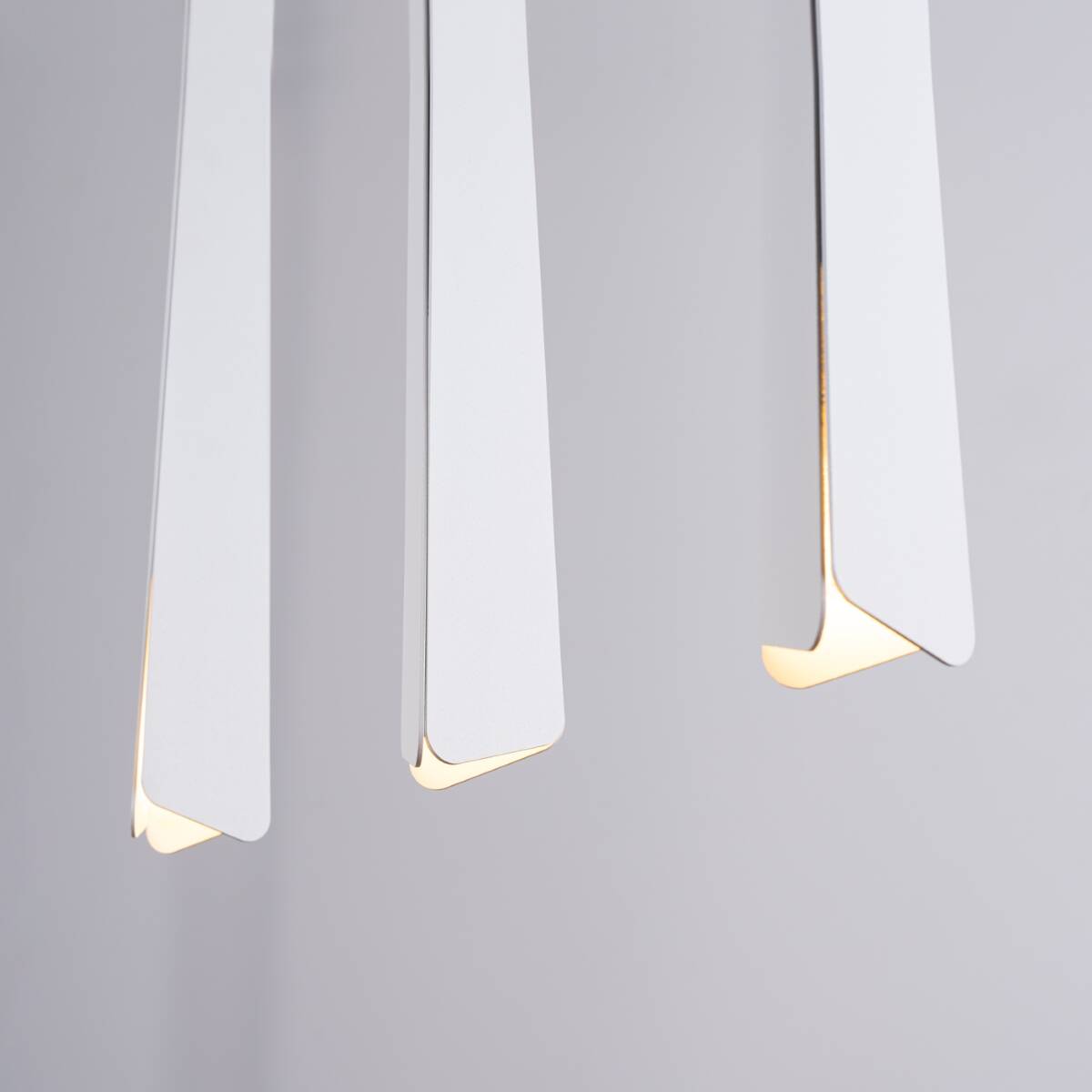Lampa wisząca Prysm 3xG9 w kolorze białym, składająca się z trzech asymetrycznych, matowych kloszy o prostokątnym kształcie, zawieszonych pod kątem. Ciepłe światło wydobywa się z ich dolnych krawędzi, na szarym tle.