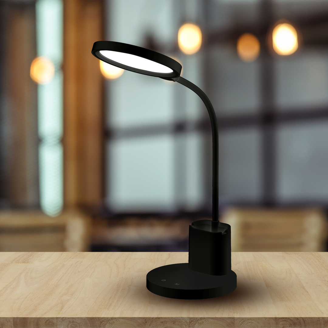 Czarna lampa biurkowa Scuola z jedną diodą LED 8W. Posiada zakrzywioną ramię i okrągłą podstawę. Widoczny przycisk włączania na podstawie. Ujęcie z boku na tle jasnego wnętrza.