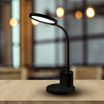 Czarna lampa biurkowa Scuola z jedną diodą LED 8W. Posiada zakrzywioną ramię i okrągłą podstawę. Widoczny przycisk włączania na podstawie. Ujęcie z boku na tle jasnego wnętrza.