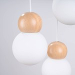 Lampa wisząca Pekka 3P z trzema źródłami światła G9, złożona z białych, okrągłych kloszy i drewnianych, okrągłych elementów u góry. Produkt zawieszony na białym przewodzie, na jasnym tle.