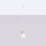 Lampa wisząca Pekka z pojedynczym źródłem światła G9, zawieszona na cienkim białym przewodzie. Posiada okrągły klosz z matowego szkła oraz drewnianą, jasną kulę u góry. Tło w odcieniach szarości, minimalistyczny design.