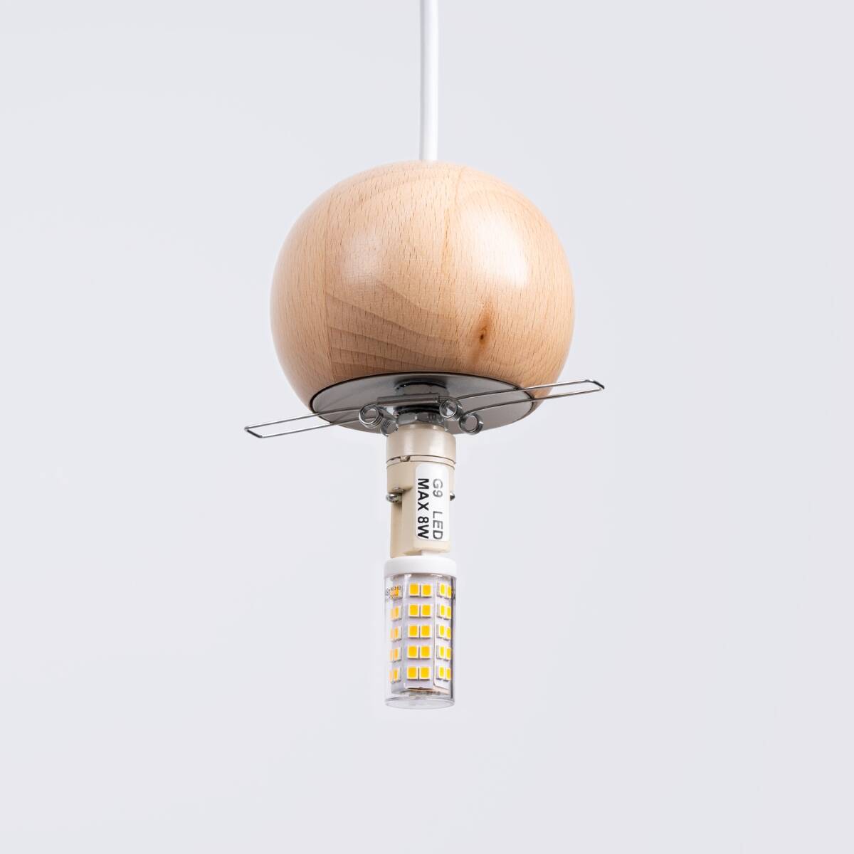 Lampa wisząca Pekka z drewnianą kulą w kolorze naturalnego drewna, z oprawką G9. Zawieszona na białym przewodzie, z widoczną żarówką LED. Kula ma gładką powierzchnię, a metalowa podstawka zabezpiecza żarówkę. Tło proste, szare.