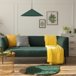 Lampa wisząca Capital D60 w butelkowej zieleni, z kloszem w kształcie stożka, zawieszona nad zieloną sofą. W tle widoczne roślinne grafiki oraz dodatki w kolorze żółtym i czarnym. Idealna do nowoczesnych aranżacji wnętrz.