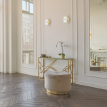 Kinkiet Elegance 1xLED/9W w złotym wykończeniu, montowany na ścianie. Oświetlenie o nowoczesnym designie, z geometrycznym kloszem, w otoczeniu eleganckiego wnętrza z drewnianą podłogą i stylowym stolikiem.