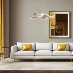 Lampa wisząca Largo 1xLED/16W w kolorze złotym, o falistym kształcie, zawieszona nad jasną sofą z poduszkami w złotym kolorze. Tło w stonowanych odcieniach, z lustrzanym obrazem i złotymi zasłonami.