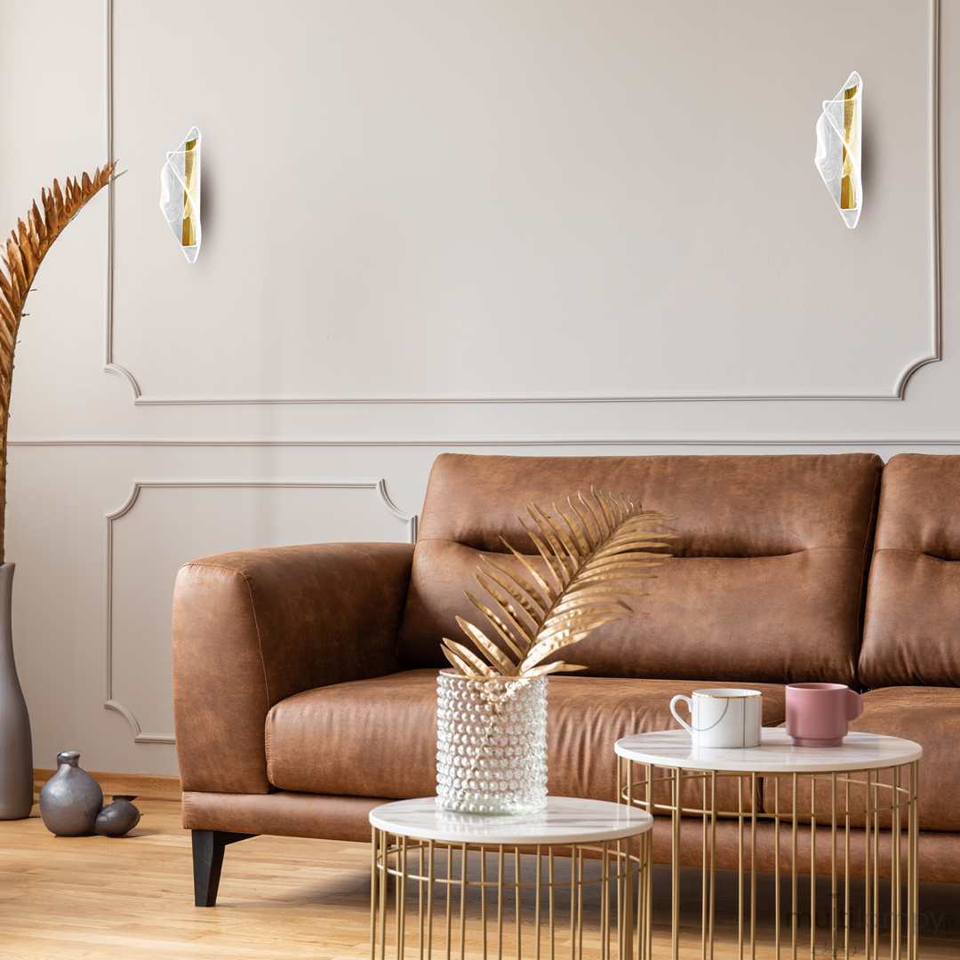 Largo kinkiet 1xLED/8W w złotym kolorze, w formie owalnej, zamontowany na ścianie. W tle widoczna brązowa sofa, stoliki kawowe oraz dekoracje roślinne, na jasnym tle.