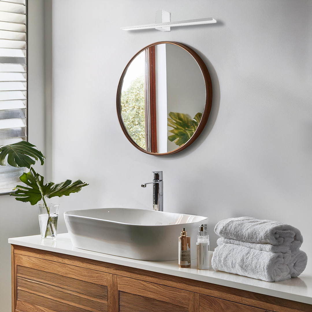 Kinkiet LED Artemis w kolorze chromu, o wymiarach 60 cm i mocy 13,8W, zamontowany nad okrągłym lustrem. Produkt o temperaturze barwowej 4000K, umieszczony w nowoczesnej łazience z drewnianą szafką, umywalką i roślinnością.