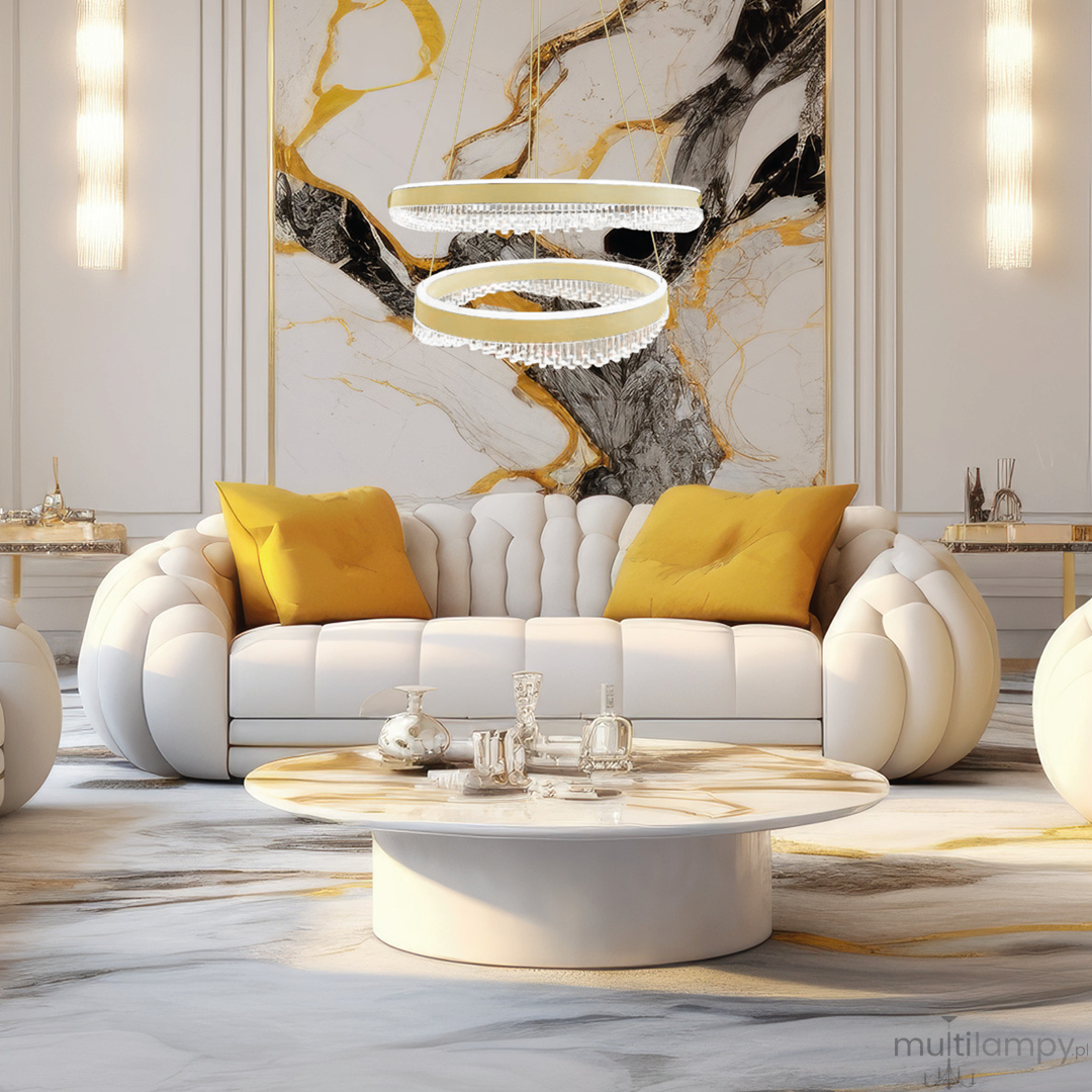 Lampa wisząca Preciosa z dwiema oprawami LED, w kolorze złotym, z przezroczystymi kryształkami. Zawieszona nad okrągłym stołem w nowoczesnym wnętrzu, z białą sofą i żółtymi poduszkami.