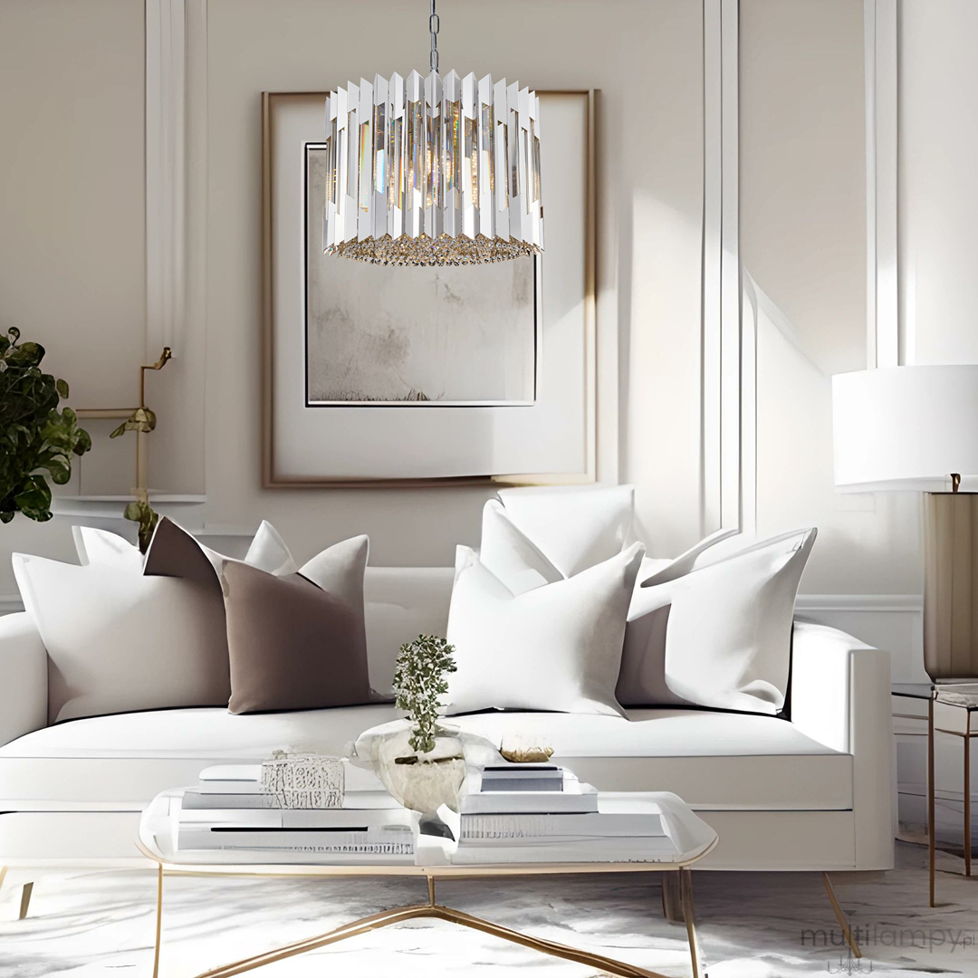 Lampa wisząca Ritz 7xE14 w chromowanej oprawie z dekoracyjnymi elementami, zawieszona nad białą sofą z poduszkami w odcieniach szarości. Jasne, nowoczesne wnętrze z minimalistycznymi dodatkami i dużą ilością naturalnego światła.