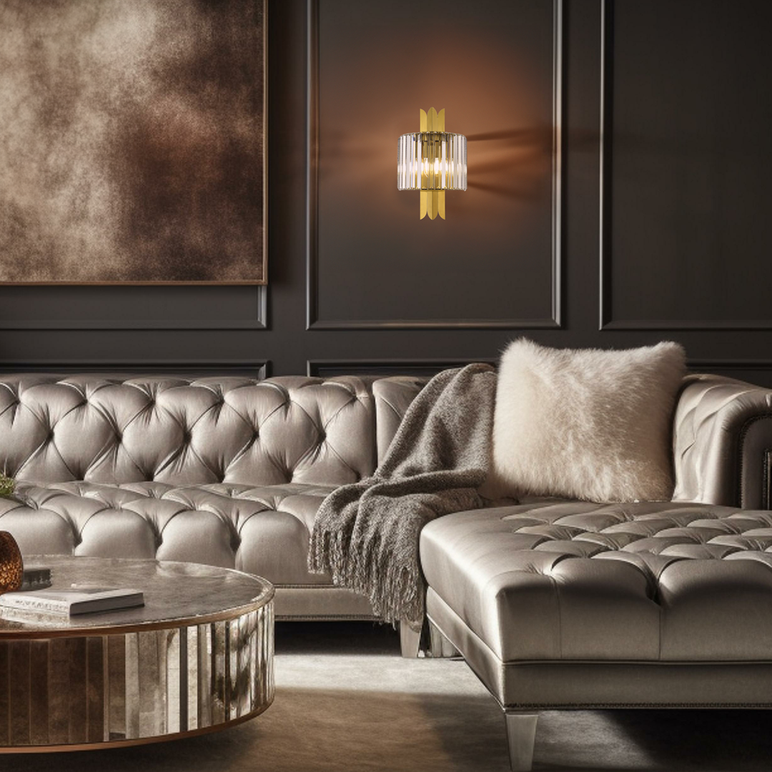 Kinkiet Margot 2xE14 w kolorze złotym, z przezroczystymi kloszami, zamontowany na ciemnej ścianie. Obok elegancka, tapicerowana sofa w odcieniu srebrnym oraz okrągły stół z lustrzanym wykończeniem.