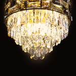 Lampa sufitowa Luxuria 9xE14 w złotej oprawie, z kryształowymi elementami w kształcie łezki, emitująca jasne światło. Ujęcie z dołu, ukazujące detale dekoracyjne oraz blask kryształów na ciemnym tle.