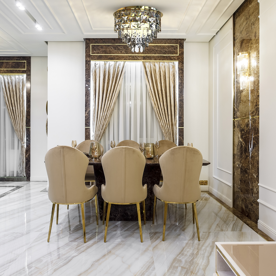 Lampa sufitowa Luxuria 9xE14 w kolorze złotym, zawieszona na suficie. Ma kryształowy klosz z błyszczącymi detalami, umieszczona w eleganckim wnętrzu z marmurowymi akcentami i miękkimi, beżowymi krzesłami wokół stołu.