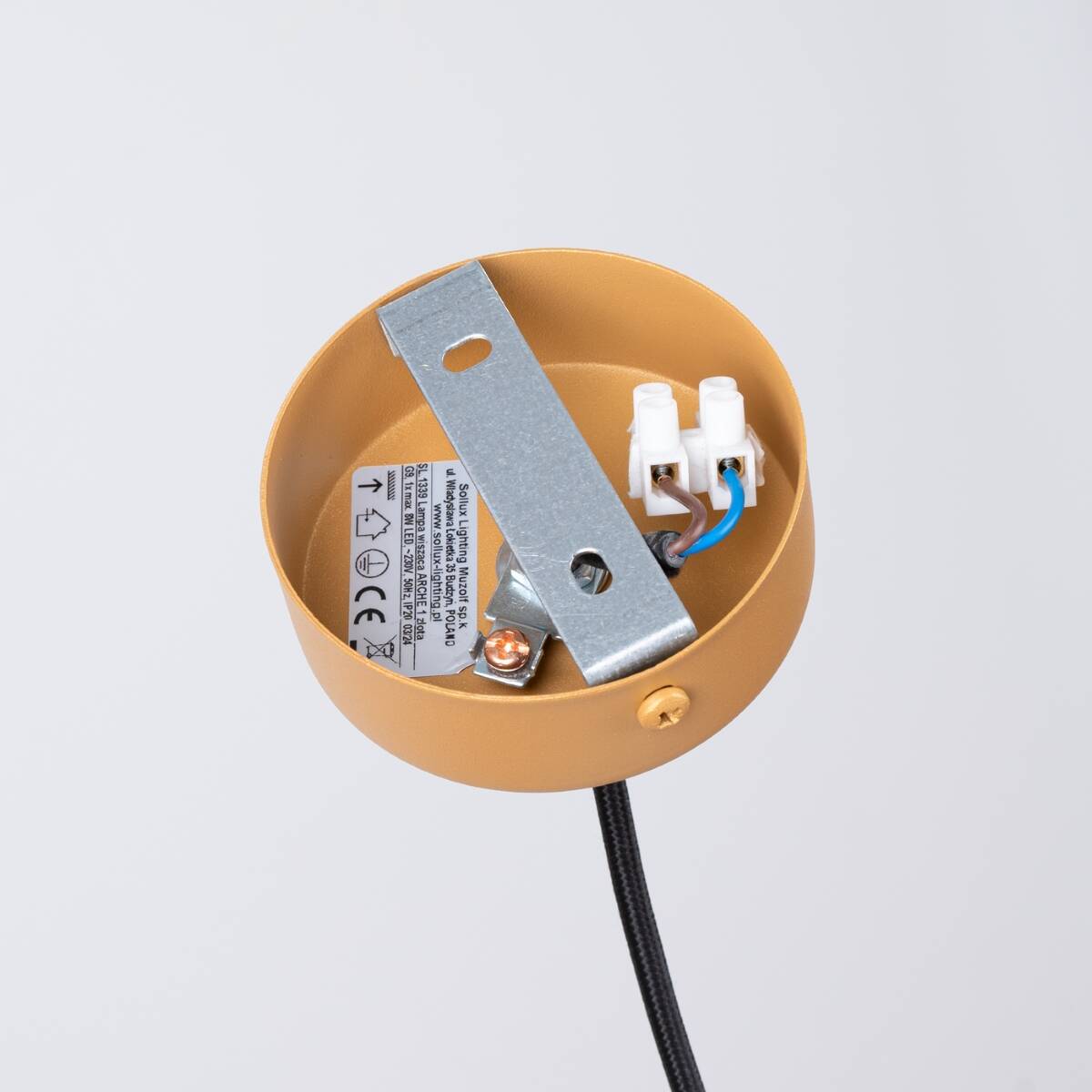 Złota lampa wisząca Arche z jednym gniazdem G9, widok od góry. Metalowa osłona z oznaczeniami technicznymi, uchwyt montażowy ze stalowym elementem. Przewód elektryczny czarny. Tło jasne, proste.