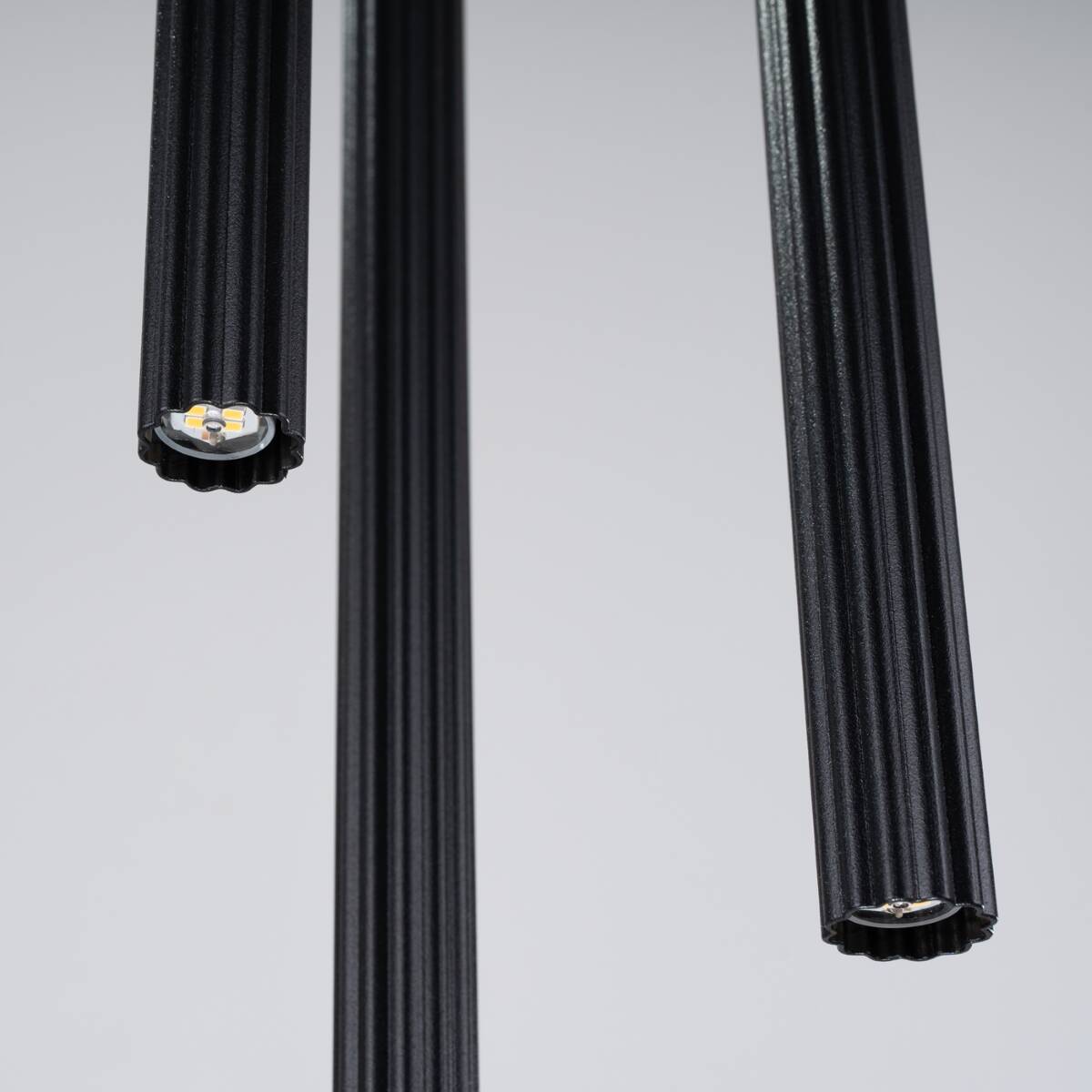 Lampa wisząca Arche w kolorze czarnym, z trzema cylindrycznymi oprawami na żarówki G9. Widoczne teksturowane detale i wewnętrzne diody LED. Ujęcie z bliska, ukazujące końcówki lampy. Proste, jasne tło.