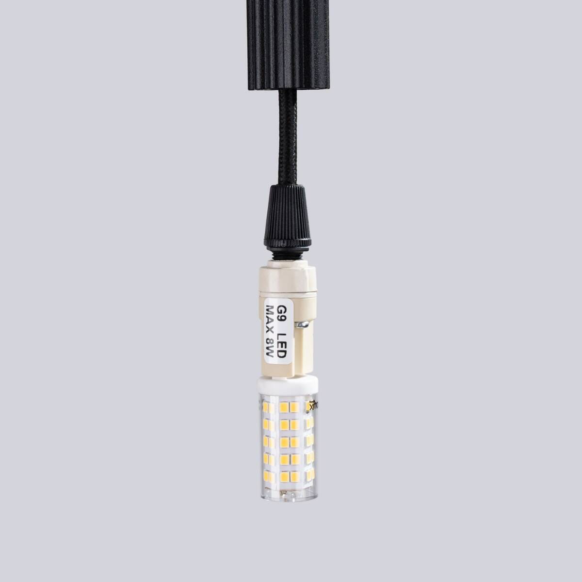 Lampa wisząca Arche w kolorze czarnym z trzema gniazdami G9. Widoczny detal to żarówka LED w osłonie szklanej, zawieszona na czarnym kablu. Kąt ujęcia zbliżony, pokazujący gniazdo i źródło światła. Tło w odcieniu jasnoszarym.