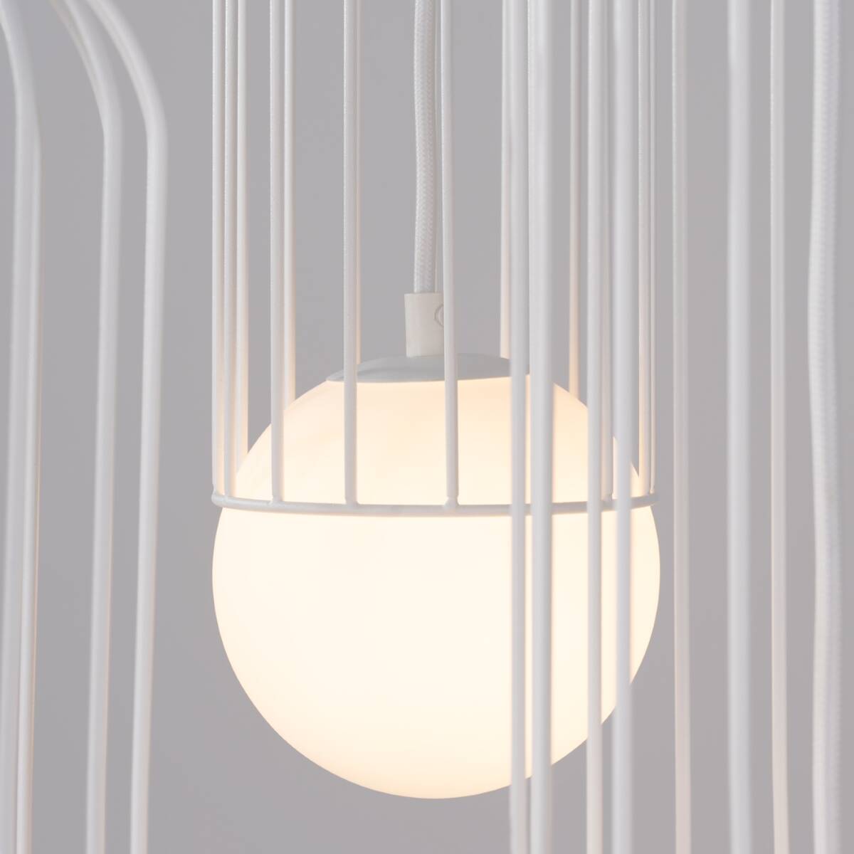 Lampa wisząca Brina 3P w kolorze białym z trzema źródłami światła G9. Widoczna okrągła, matowa kula w metalowej, białej klatce. Ujęcie z bliska, koncentrujące się na detalu lampy i jej struktury.