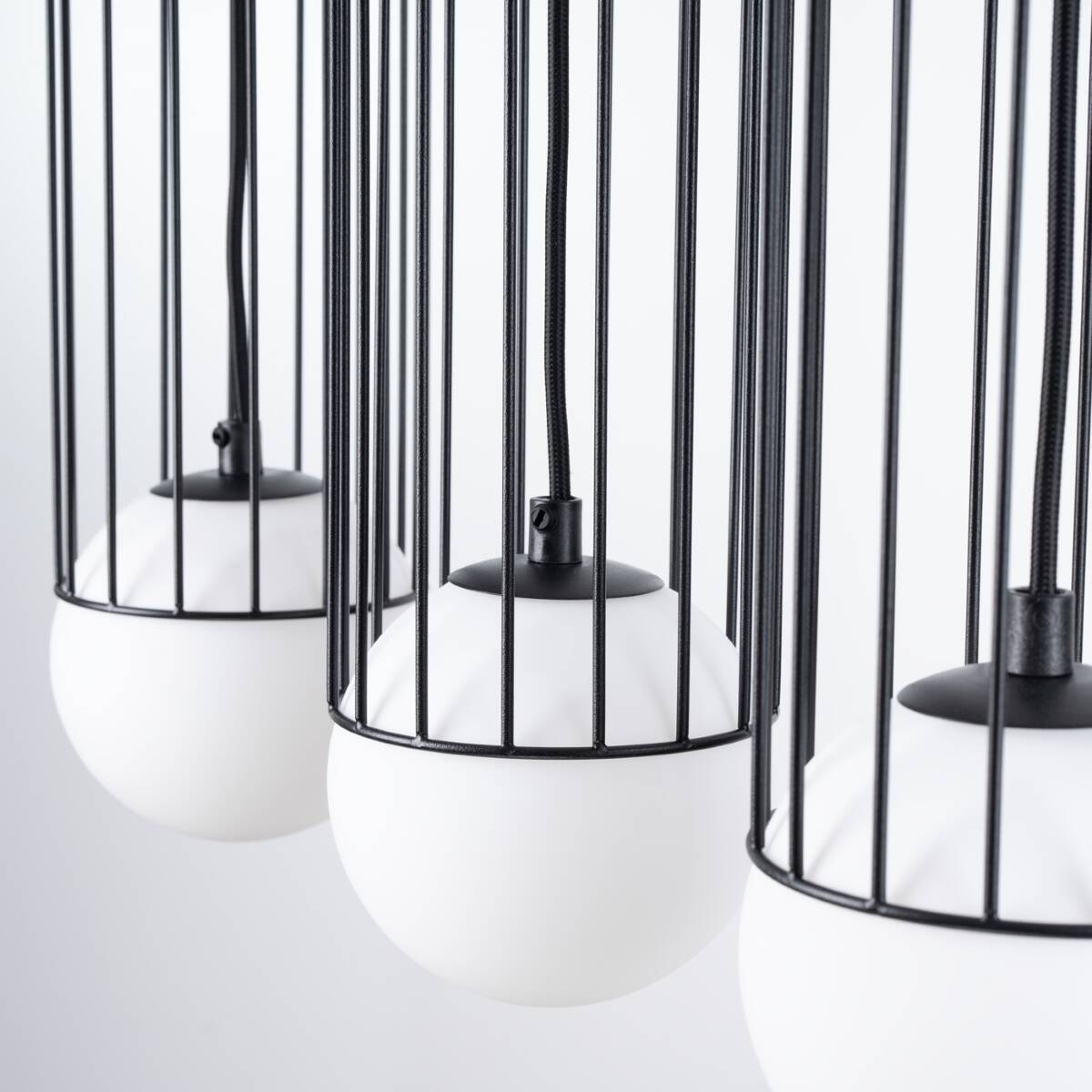 Lampa wisząca Brina 3xG9 w kolorze czarnym, z trzema białymi kloszami w kształcie kul, osadzonymi w metalowej siatce. Kable zawieszenia w czarnym kolorze. Widok z bliska, ukazujący detal konstrukcji.