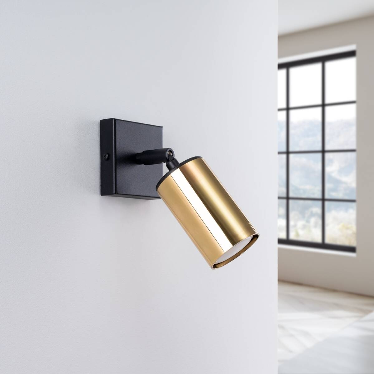 Kinkiet Winx w kolorze czarnym i złotym, z cylindryczną oprawą na żarówkę GU10. Ujęcie z boku, zamontowany na ścianie, z widoczną złotą częścią reflektora. Tło jasne, z dużym oknem w tle.
